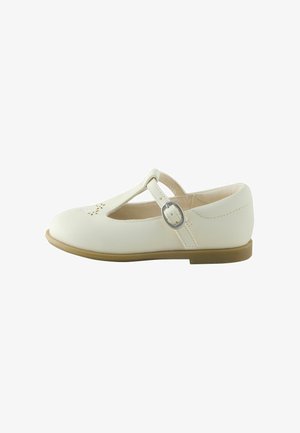 White leather Mary Jane shoe with a rounded toe, T-strap design, ankle buckle, and cut-out flower pattern on the toe. Flat tan sole.
