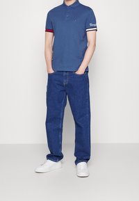 Blå polo med kontrasterande röda och vita randiga ärmar, i kombination med löst sittande mörkblå jeans och vita sneakers.