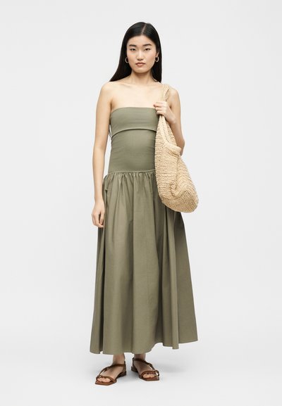 Γυναίκα που φοράει στράπλες μακρύ φόρεμα σε ελαιδί πράσινο χρώμα, καφέ φλατ σανδάλια, κρατώντας μεγάλη μπεζ υφαντή τσάντα ώμου, στέκεται μπροστά σε απλό φόντο.