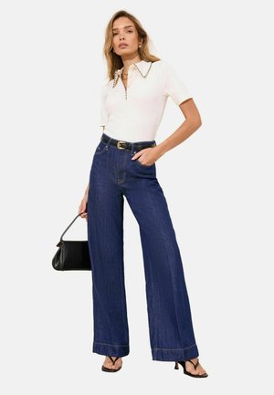 Kobieta ubrana w biały polo z krótkim rękawem z czarnym obszyciem, ciemnoniebieskie dżinsy o wysokim stanie i szerokich nogawkach, czarny pasek, czarną torebkę i czarne sandały na obcasie.
