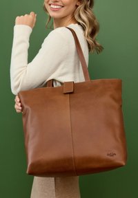 Naine, kes kannab kreemikat kampsunit, hoiab suurpruunist nahast kaherihmalist käekotti rohelise tausta ees.