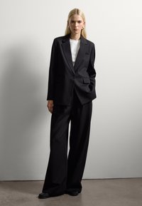 Completo nero su misura caratterizzato da un blazer strutturato con un bottone singolo e pantaloni a gamba larga, realizzato in tessuto liscio. Sottostante una maglietta bianca.