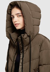 Noor naine kannab pruunist pufferjacketit, millel on suur kapuuts ja mis on osaliselt tõmmatud, halli ühetoonilise tausta ees.