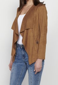 Femme portant une veste en daim marron ouverte sur le devant avec col drapé, un haut blanc et un jean bleu taille haute sur un fond blanc uni.