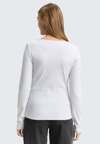 Witte langemouwen top met een ribbelstructuur en een brede halslijn, gecombineerd met donkergrijze broek, gezien vanuit de achterkant.