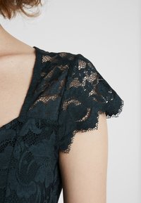 Tmavě modrozelené krajkové šaty s propracovanými květinovými vzory, volánovými rukávy a texturovanou vrstvou. Podkladem je hladká látka pro pohodlí.