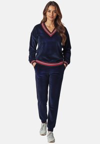 Marineblauer Velours-Tracksuit mit V-Ausschnitt, ausgestattet mit roten und weißen gestreiften Akzenten an den Bündchen. Die Hose hat eine lockere Passform und seitliche Taschen.