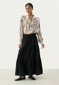 Blusa con stampa paisley in bianco e nero, colletto annodato; abbinata a una lunga gonna nera a strati. Tessuto morbido; il modello indossa scarpe nere.