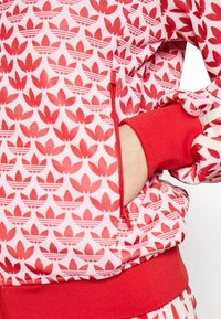 Röd jacka med dragkedja och ett repetitivt bladmönster i varierande nyanser av röd och rosa. Har röda ribbade ärmslut och nederkant.