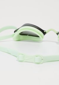 Hellgrüne Schwimmbrille mit schwarzen Gläsern, die auf einer weißen Oberfläche liegt und die verstellbaren Riemen und Detail des Clips zeigt.