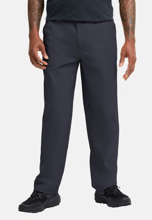 Uomo che indossa pantaloni dritti blu scuro e scarpe sportive nere, in piedi con le braccia rilassate lungo i fianchi su sfondo bianco.
