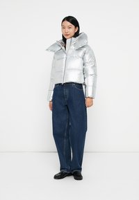 Silver puffer jacket with wide collar, zip front, and glossy texture, paired with loose-fitting dark blue jeans and black shoes.
