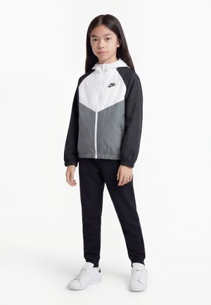 Młoda osoba stojąca w kolorowej kurtce Nike, czarnych spodniach i białych sneakersach na jednolitym białym tle.