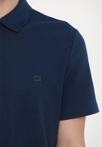 Jūrinės mėlynos spalvos polo marškiniai su tekstūruota medžiaga, trumpomis rankovėmis ir mažą juodą logotipą kairėje krūtinės pusėje. Klasikinio tipo apykaklė.