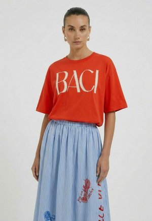 Woman in red oversized T-shirt with "BACI" text and blue striped skirt with red and blue embroidery, standing against plain background.