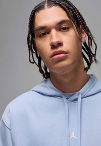 Hoodie azul-claro com um bolso frontal, cordão e logo Jumpman branco no peito. O modelo tem cabelo trançado e uma expressão neutra.