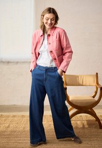 Femme souriante les mains dans les poches, portant une veste rose, un chemisier blanc, un pantalon large bleu, debout à côté d'une chaise en bois sur un tapis tissé.