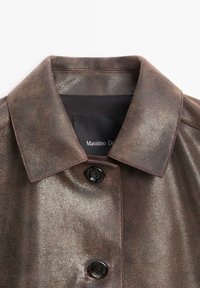 Brun jakke af imiteret læder med en glat tekstur, en klassisk krave og to sorte knapper foran. Logomærke indeni kraven.