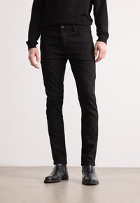 Jeans slim-fit noirs au toucher lisse, avec un design classique à cinq poches et des coutures discrètes, associés à des bottes en cuir noires à la cheville.