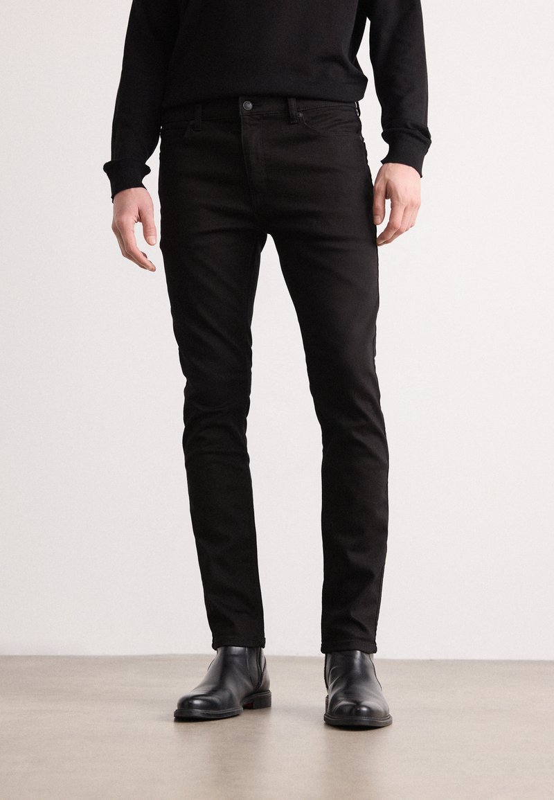 Jeans slim-fit noirs au toucher lisse, avec un design classique à cinq poches et des coutures discrètes, associés à des bottes en cuir noires à la cheville.