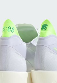 Baskets blanches avec languettes de traction vert néon, chiffres de style numérique sur la languette gauche, et symbole géométrique sur la languette droite, vues de dos.