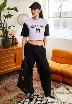 T-shirt noir et lavande coupée avec l'imprimé "NEW YORK", pantalon noir ample et casquette noire. Tenue avec un sac fourre-tout noir matelassé.