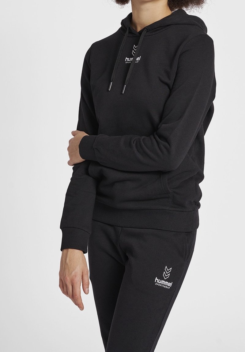 Personne portant un hoodie de sport Hummel noir et un pantalon de survêtement noir assorti, bras croisés devant un fond uni.