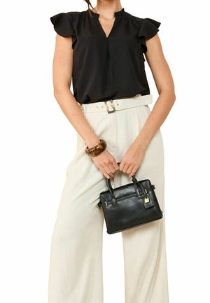 Femme portant un chemisier noir à manches courtes et un pantalon blanc taille haute, tenant un petit sac à main noir avec un bracelet marron au poignet gauche.