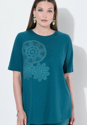 Kobieta w morskim topie z krótkim rękawem z koronkowymi kwiatowymi wzorami z przodu, długimi prostymi włosami i dużymi wiszącymi kolczykami na białym tle.