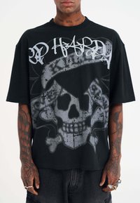 Camiseta de gran tamaño en negro con un gráfico de una calavera gris con huesos cruzados y texto en estilo graffiti en la parte frontal. Material de algodón, mangas cortas.