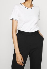 Camiseta blanca con ribetes negros en el cuello y las mangas, combinada con pantalones negros ajustados. La camiseta tiene un corte relajado y una textura suave.
