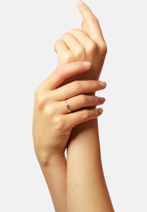 Goldring mit einem zentralen Diamanten, getragen an einer Hand mit glatter Haut und neutralem Nagellack, die Finger miteinander verwoben vor einem hellen Hintergrund.