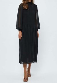 Černé šaty s dlouhým rukávem z průhledné látky se stahovaným pasem. Mají vysoký výstřih a nafouklé rukávy, doplněné o hnědé podpatky.