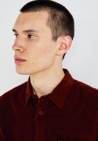 Camisa de pana marrón con cuello abotonado, que cuenta con un bolsillo con un pequeño logotipo, usada por una persona con el cabello corto sobre un fondo neutro.