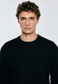 Giovane uomo con capelli ricci fissa direttamente la fotocamera, indossando un maglione nero a trama su uno sfondo chiaro e uniforme.