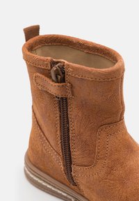 Bota de cuero marrón con detalle de grabado floral, cierre de cremallera lateral y suela cosida sobre un fondo claro.