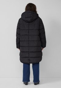 Schwarze Pufferjacke mit Kapuze, mit horizontal gesteppten Abschnitten, die bis unter die Knie reicht, kombiniert mit blauen weit geschnittenen Jeans.