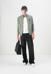 Light green zip-up jacket over a white t-shirt, black wide-leg jeans, white sneakers, and a black cap. Holding a black tote bag.