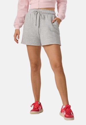 Femme portant un short gris à cordon, un sweat à capuche court rose et des baskets rouge vif, debout avec une main dans la poche.