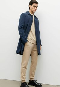 Granatowy wodoodporny płaszcz trench z kołnierzem i zapięciem na guziki, noszony na beżowej bluzie i dopasowanych dresowych spodniach; czarne buty na sznurowadła.