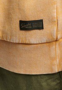 Oranžová texturovaná látka s žebrovaným lemem a manžetami. Obsahuje černou etiketu s bílým textem uvádějícím značku a výrobní detaily.