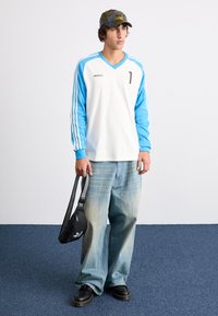 Chemise blanche à manches longues avec des manches bleues, arborant le logo Adidas et le numéro 1. Associée à un jean large délavé et un sac bandoulière noir.