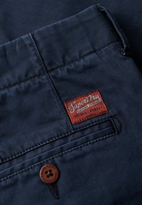 Marineblauwe stof met een gladde textuur, voorzien van een bruine Superdry-label en een bruine knoopdetail op de zak.