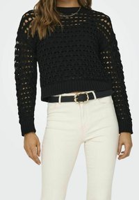 Schwarzer Häkelpullover mit offenem Maschenmuster, rundem Ausschnitt und gerippten Bündchen. Kombiniert mit weißen High-Waist-Jeans und einem schwarzen Gürtel.