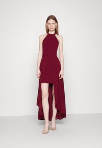 Burgundy halter dress with a fitted bodice and asymmetric hem, featuring a flowing skirt and soft texture. Silver strappy heels accent the look.
