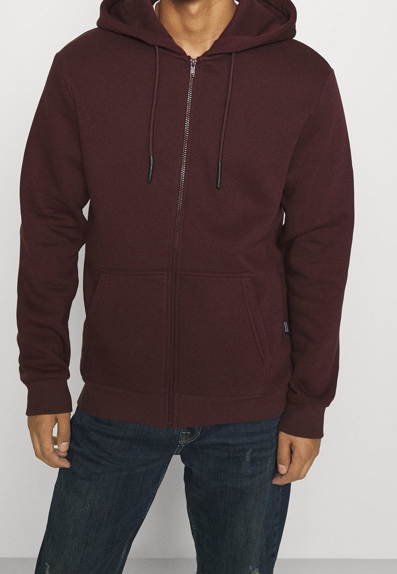 Homme portant un sweat à capuche zippé rouge foncé avec des poches avant et un jean bleu, debout devant un fond clair uni.