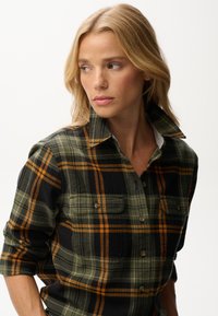 Green and black plaid flannel shirt with orange accents, featuring a collared neckline and two front pockets, textured fabric.
