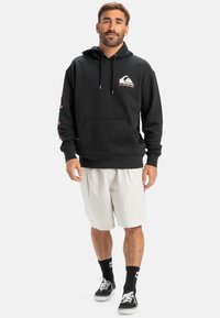 Black hooded sweatshirt with front pocket, drawstrings, and logo. Features print details on sleeves. Paired with light shorts and socks.