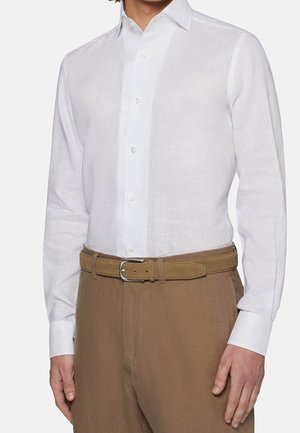 Mies, joka käyttää valkoista pitkähihasta napillista paitaa, joka on tungettu ruskeisiin housuihin ja ruskeaan vyöhön.