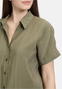 Olijfgroen overhemd met korte mouwen, een kraag, knoopsluiting en een gladde, licht gekreukte stofstructuur.